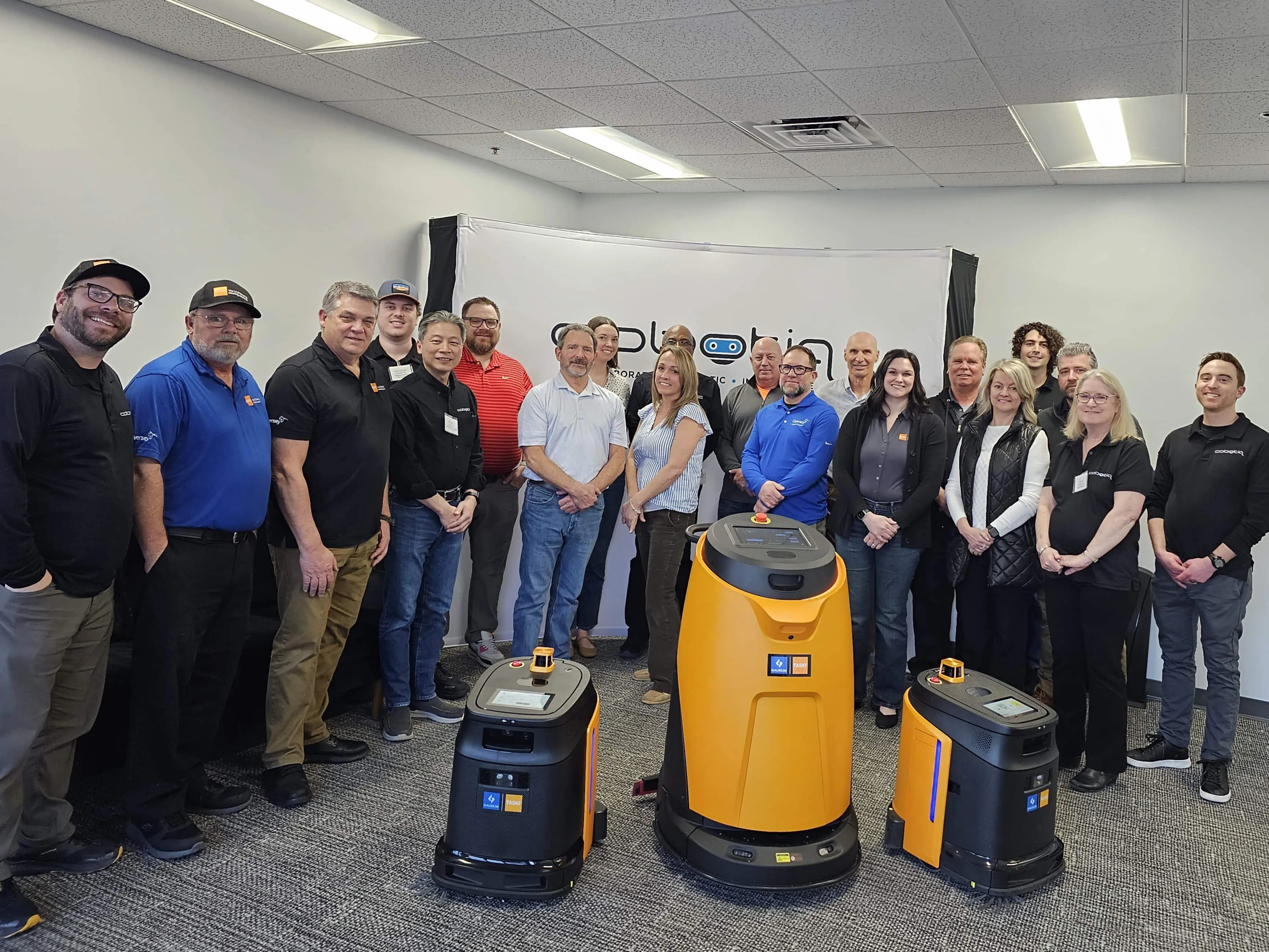 The Cobotiq team standing with three autonomous cleaning robots in front of the Cobotiq banner